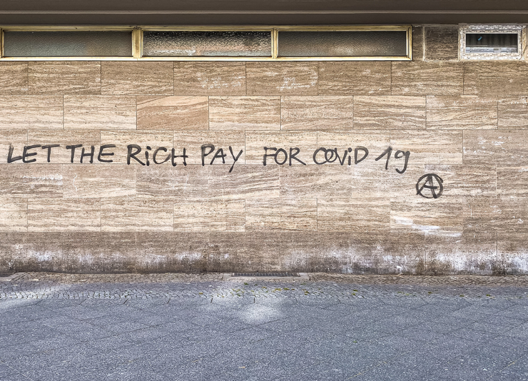 Kuvassa vaaleanruskea tiiliseinä, johon on kirjoitettu teksti: Let the rich pay for covid 19.