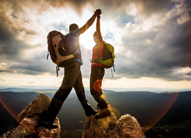 Kuvassa näkyy mies ja nainen, molemmat ovat vuoren huipulla ja pitävät toisiaan kädestä kiinni. Kädet ovat ylhäällä kohotettuna kohti taustalla näkyvää auringonvaloa. Taustalla näkyy myös maisema alas laaksoon.