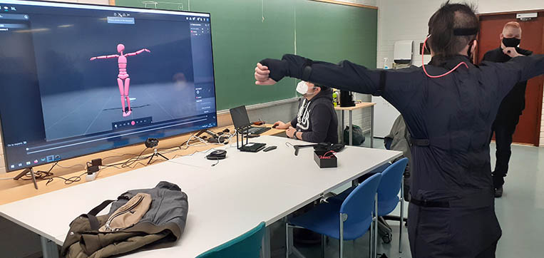 Kuvassa näkyy henkilö, jollan on päällään liikkeentunnistusteknologiaan liittyvä puku. Vieressä on iso näyttö, jossa henkilön liike toistuu mallinnettuna tietokoneella. Molemmat hahmot ovat samassa asennossa.