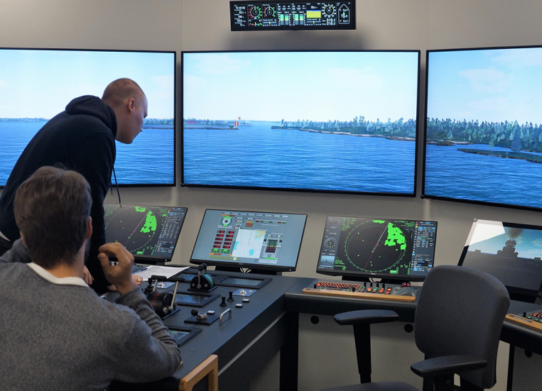 Kaksi merikapteeniopiskelijaa ohjaa laivaa simulaattorissa kohti Suomenlinnan ja Vallisaaren välissä olevaa Kustaanmiekan salmea Helsingissä. Isoista monitoreista näkymä on verrattavissa aluksen tutkien tuottamaan kuvaan konsolitasojen näytöissä.
