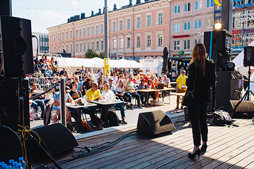 In the front of the picture there is a host on the stage, behind her is seen people listening in the marketplace.