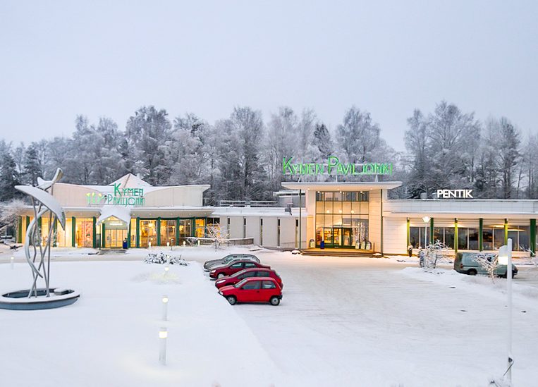 The Kymen Paviljonki -building is photographed outside in winter. There is lots of snow around, and a few cars in front of the building, with a frosty forest in the background.