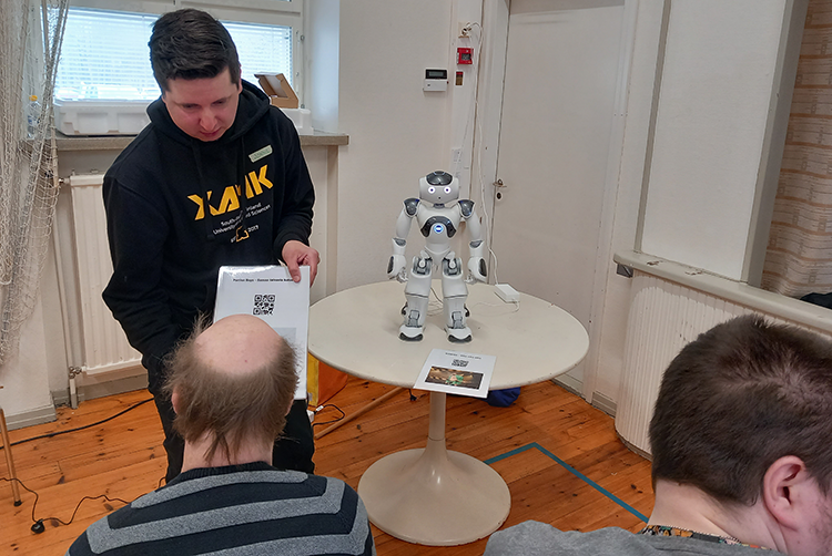 Zora-robotti seisoo pöydällä odottamassa seuraavaa komentoa. Robotin ohjaamista opastamassa hankkeen henkilökunta ja oppimassa Sotek-säätiön asiakas.