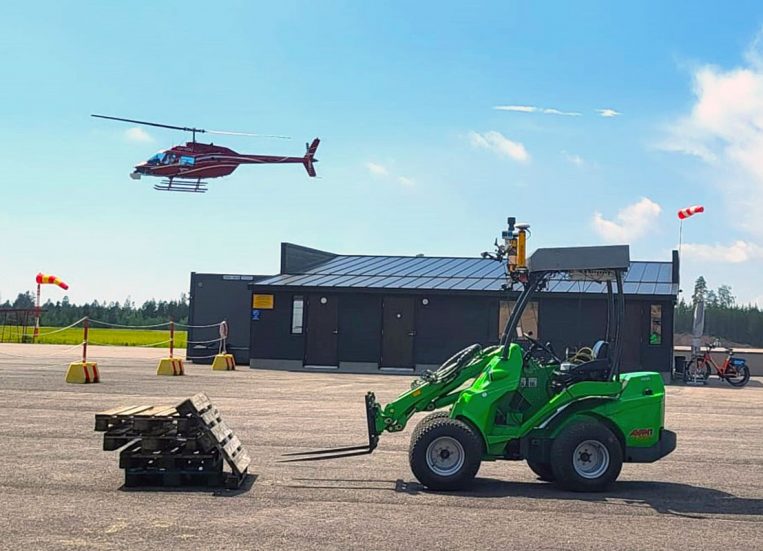 Kuvassa vihreä työkone harmaan rakennuksen edessä. Koneen edessä on pino kuormalavoja. Näiden yllä ohitse lentää punainen helikopteri sininen taivas taustanaan. Taustalla näkyy metsän raja ja työkoneen ympäristö on rajattu betoniporsain.